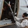 Green Berets, Danish SOF Ski Across Greenland Ice Cap During Operation Apex Indigo A person in a white winter suit and tactical vest is loading or unloading ski equipment into/from a military-style helicopter. The individual's face has been deliberately blurred, suggesting operational security or privacy concerns. The scene appears to take place on a tarmac or airfield in an overcast environment.