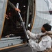 A person in a white winter suit and tactical vest is loading or unloading ski equipment into/from a military-style helicopter. The individual's face has been deliberately blurred, suggesting operational security or privacy concerns. The scene appears to take place on a tarmac or airfield in an overcast environment.