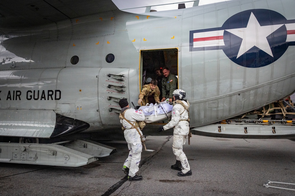 Personnel in white protective suits and military uniforms are transferring a patient on a stretcher through the side door of a US Air National Guard military transport aircraft. The operation appears to be a medical evacuation or MEDEVAC exercise, with at least four individuals involved in the transfer. The aircraft bears the US military roundel insignia and partial text reading 'AIR GUARD' on its fuselage.