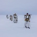 A column of military personnel in white winter camouflage uniforms are cross-country skiing across a flat, snow-covered terrain under an overcast sky. They are carrying rifles slung across their backs and using ski poles, moving in a single-file formation. The scene appears to be an arctic or subarctic military training exercise.