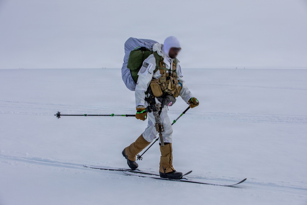 A heavily equipped military or special operations personnel is cross-country skiing across a snow-covered arctic or winter terrain. The individual is wearing white winter camouflage over tactical gear, carrying a large rucksack, and is armed with a weapon secured to their kit. The environment is flat, overcast, and snow-covered with minimal visibility in the background.