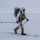 Green Berets, Danish SOF Ski Across Greenland Ice Cap During Operation Apex Indigo A heavily equipped military or special operations personnel is cross-country skiing across a snow-covered arctic or winter terrain. The individual is wearing white winter camouflage over tactical gear, carrying a large rucksack, and is armed with a weapon secured to their kit. The environment is flat, overcast, and snow-covered with minimal visibility in the background.