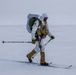 A heavily equipped military or special operations personnel is cross-country skiing across a snow-covered arctic or winter terrain. The individual is wearing white winter camouflage over tactical gear, carrying a large rucksack, and is armed with a weapon secured to their kit. The environment is flat, overcast, and snow-covered with minimal visibility in the background.