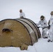 Three military personnel in winter camouflage uniforms are setting up a large tan/khaki tunnel-style tent in snowy, blizzard-like conditions. At least one individual displays a Danish flag patch on their sleeve, suggesting this is a Danish military or NATO cold-weather training exercise. Skis are visible in the background, indicating the team traveled to this location on ski.