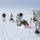 Green Berets, Danish SOF Ski Across Greenland Ice Cap During Operation Apex Indigo A group of four military personnel in white winter camouflage uniforms are skiing across a vast, flat snow-covered terrain. They are carrying large backpacks and pulling loaded pulk sleds behind them using ski poles for propulsion. The scene appears to be an arctic or subarctic military training or patrol exercise.