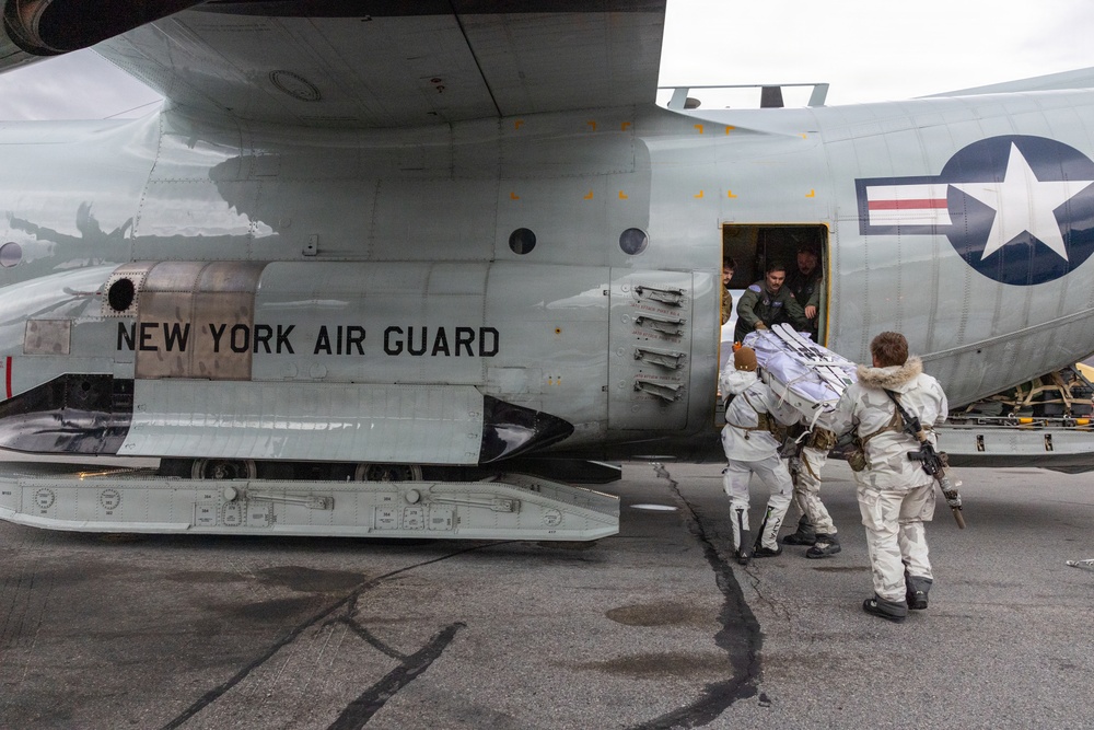 Personnel in white winter camouflage uniforms are loading or transferring cargo/equipment into a New York Air National Guard military transport aircraft. The aircraft displays the US military roundel insignia and 'NEW YORK AIR GUARD' markings on its fuselage. Crew members are visible in the open side door of the aircraft assisting with the operation.