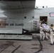 Personnel in white winter camouflage uniforms are loading or transferring cargo/equipment into a New York Air National Guard military transport aircraft. The aircraft displays the US military roundel insignia and 'NEW YORK AIR GUARD' markings on its fuselage. Crew members are visible in the open side door of the aircraft assisting with the operation.