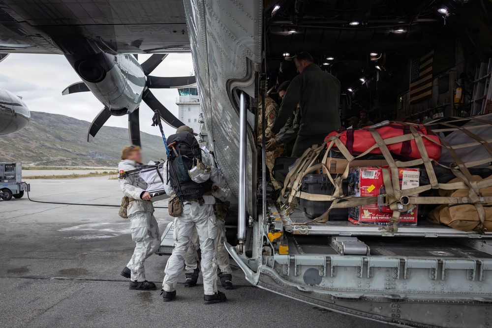 Military personnel in white cold-weather suits are boarding or loading a large military transport aircraft, likely a C-130 or similar tactical airlifter, on a tarmac with mountainous terrain visible in the background. The aircraft's rear cargo ramp is open and loaded with netted cargo including red bags, cases, and boxes of supplies including what appear to be portable heaters. An American flag is visible inside the aircraft cabin.