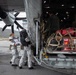 Military personnel in white cold-weather suits are boarding or loading a large military transport aircraft, likely a C-130 or similar tactical airlifter, on a tarmac with mountainous terrain visible in the background. The aircraft's rear cargo ramp is open and loaded with netted cargo including red bags, cases, and boxes of supplies including what appear to be portable heaters. An American flag is visible inside the aircraft cabin.