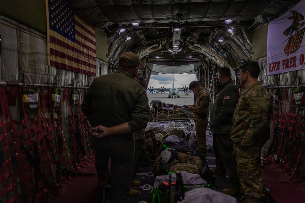 The image shows the interior of a military transport aircraft, likely a C-130 Hercules, with several personnel in military uniforms preparing equipment. An American flag is mounted on the left interior wall, and a banner reading 'LIVE FREE OR...' is visible on the right. Through the open rear cargo ramp, another C-130 aircraft is visible on the tarmac outside.