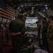 The image shows the interior of a military transport aircraft, likely a C-130 Hercules, with several personnel in military uniforms preparing equipment. An American flag is mounted on the left interior wall, and a banner reading 'LIVE FREE OR...' is visible on the right. Through the open rear cargo ramp, another C-130 aircraft is visible on the tarmac outside.