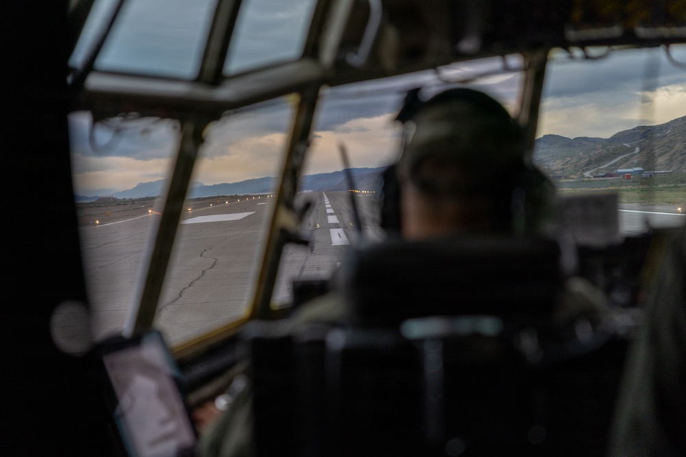 The image is taken from inside a military aircraft cockpit, showing a pilot or crew member wearing a flight helmet seated at the controls. The view through the multi-pane cockpit windows reveals a runway with edge lighting stretching into the distance, surrounded by mountainous terrain under a partly cloudy sky at dusk or dawn. The aircraft appears to be on approach or taxiing on the runway.