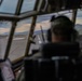 The image is taken from inside a military aircraft cockpit, showing a pilot or crew member wearing a flight helmet seated at the controls. The view through the multi-pane cockpit windows reveals a runway with edge lighting stretching into the distance, surrounded by mountainous terrain under a partly cloudy sky at dusk or dawn. The aircraft appears to be on approach or taxiing on the runway.