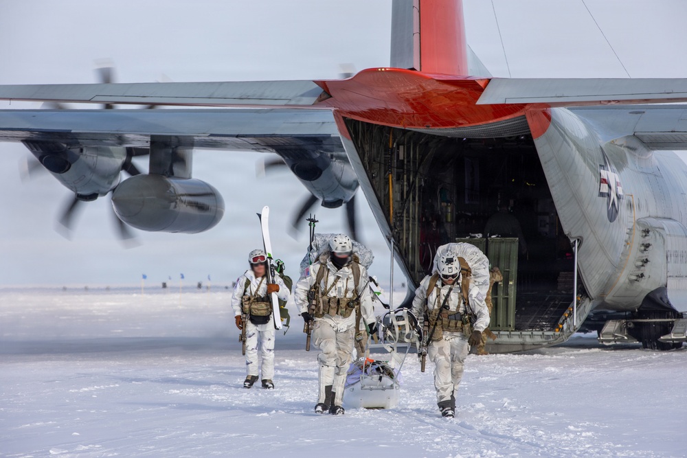 Three military personnel in white winter camouflage gear are exiting or departing from a large military transport aircraft, identified as a C-130 Hercules with a distinctive red tail section, on a snow-covered surface. The soldiers are heavily equipped with tactical gear, backpacks, weapons, and one carries skis, suggesting an arctic or cold-weather special operations mission. The aircraft engines appear to be running based on the motion blur visible on the propellers.