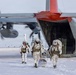 Three military personnel in white winter camouflage gear are exiting or departing from a large military transport aircraft, identified as a C-130 Hercules with a distinctive red tail section, on a snow-covered surface. The soldiers are heavily equipped with tactical gear, backpacks, weapons, and one carries skis, suggesting an arctic or cold-weather special operations mission. The aircraft engines appear to be running based on the motion blur visible on the propellers.