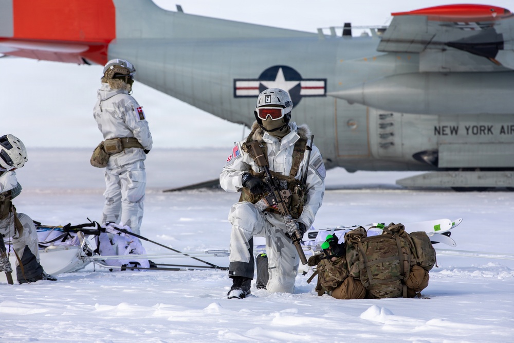Military personnel in white winter camouflage gear are conducting cold-weather operations on a snow-covered surface near a U.S. Air National Guard aircraft. The aircraft bears the inscription 'NEW YORK AIR' and displays a U.S. Air Force roundel insignia. Soldiers are equipped with weapons, skis, sleds, and tactical backpacks, suggesting an Arctic or winter warfare training exercise.