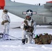 Military personnel in white winter camouflage gear are conducting cold-weather operations on a snow-covered surface near a U.S. Air National Guard aircraft. The aircraft bears the inscription 'NEW YORK AIR' and displays a U.S. Air Force roundel insignia. Soldiers are equipped with weapons, skis, sleds, and tactical backpacks, suggesting an Arctic or winter warfare training exercise.