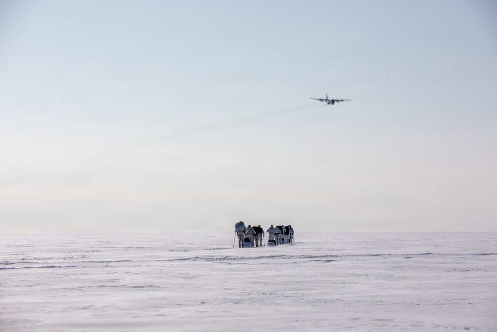 A group of personnel in white winter camouflage gear are gathered on a vast, flat snow-covered terrain, carrying equipment and sleds. A large military transport aircraft, consistent with a C-130 Hercules, flies overhead in a pale, hazy sky. The scene strongly suggests a cold-weather military training exercise or Arctic/polar operation.