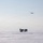 Green Berets, Danish SOF Ski Across Greenland Ice Cap During Operation Apex Indigo A group of personnel in white winter camouflage gear are gathered on a vast, flat snow-covered terrain, carrying equipment and sleds. A large military transport aircraft, consistent with a C-130 Hercules, flies overhead in a pale, hazy sky. The scene strongly suggests a cold-weather military training exercise or Arctic/polar operation.