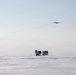 A group of personnel in white winter camouflage gear are gathered on a vast, flat snow-covered terrain, carrying equipment and sleds. A large military transport aircraft, consistent with a C-130 Hercules, flies overhead in a pale, hazy sky. The scene strongly suggests a cold-weather military training exercise or Arctic/polar operation.