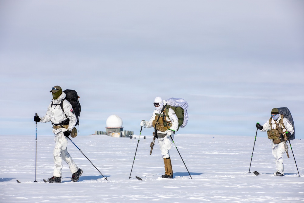 Three individuals in white winter camouflage military gear are cross-country skiing across a snow-covered arctic or polar landscape. They are carrying large backpacks and tactical vests while using ski poles, suggesting a military cold-weather training or patrol operation. A radar dome installation and what appears to be a military or research facility is visible in the background.