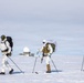 Three individuals in white winter camouflage military gear are cross-country skiing across a snow-covered arctic or polar landscape. They are carrying large backpacks and tactical vests while using ski poles, suggesting a military cold-weather training or patrol operation. A radar dome installation and what appears to be a military or research facility is visible in the background.