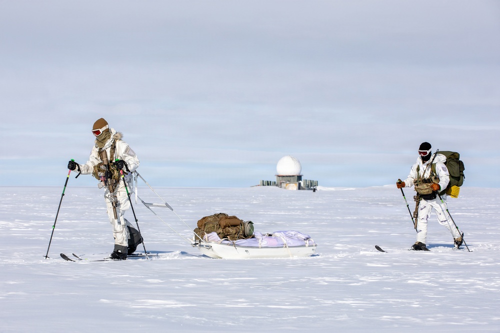 Two individuals in white winter camouflage military gear are skiing across a snow-covered arctic or polar landscape. They are equipped with weapons, tactical gear, and backpacks, and are pulling a pulk sled loaded with supplies. A radar dome installation is visible in the background, suggesting a military or research outpost.