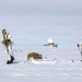 Two individuals in white winter camouflage military gear are skiing across a snow-covered arctic or polar landscape. They are equipped with weapons, tactical gear, and backpacks, and are pulling a pulk sled loaded with supplies. A radar dome installation is visible in the background, suggesting a military or research outpost.