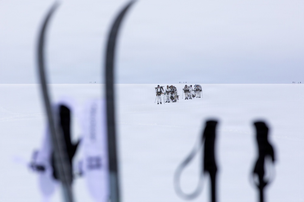 A group of approximately 6-8 people dressed in white winter gear are traversing a vast, flat snow-covered landscape in the distance. The foreground features out-of-focus ski equipment including skis and poles in silhouette, creating a framing effect. The scene suggests a polar or arctic expedition, with additional figures barely visible on the horizon.