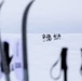 A group of approximately 6-8 people dressed in white winter gear are traversing a vast, flat snow-covered landscape in the distance. The foreground features out-of-focus ski equipment including skis and poles in silhouette, creating a framing effect. The scene suggests a polar or arctic expedition, with additional figures barely visible on the horizon.