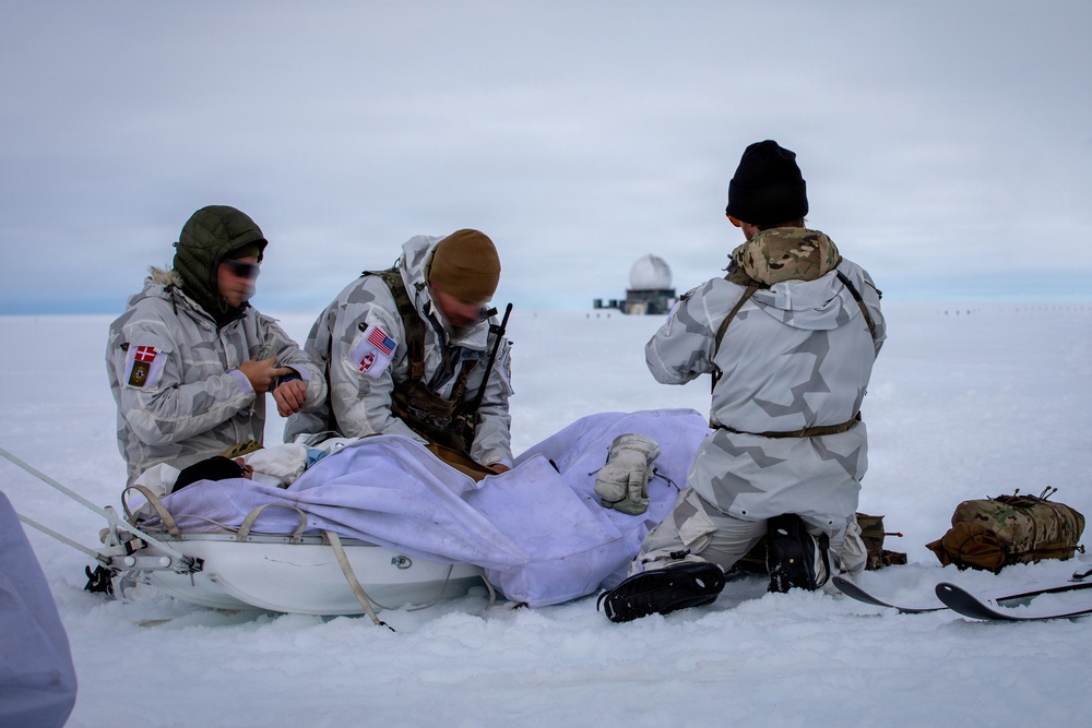Three military personnel in white winter camouflage uniforms are conducting what appears to be a field medical training exercise on a snow-covered arctic terrain. They are attending to a simulated casualty lying on a white rescue sled (pulk/ahkio). A radar dome installation is visible in the background, suggesting proximity to a military or research outpost.