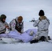 Three military personnel in white winter camouflage uniforms are conducting what appears to be a field medical training exercise on a snow-covered arctic terrain. They are attending to a simulated casualty lying on a white rescue sled (pulk/ahkio). A radar dome installation is visible in the background, suggesting proximity to a military or research outpost.