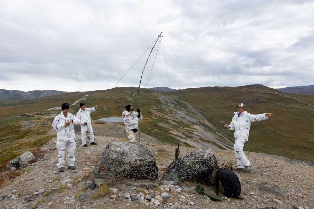Four individuals wearing white camouflage overgarments are gathered on a rocky hilltop or ridge in a mountainous, tundra-like landscape. They appear to be setting up or operating a field radio antenna mast, with guy-wires extending outward from a central pole. The terrain, vegetation, and overcast sky suggest a subarctic or alpine environment.