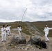 Four individuals wearing white camouflage overgarments are gathered on a rocky hilltop or ridge in a mountainous, tundra-like landscape. They appear to be setting up or operating a field radio antenna mast, with guy-wires extending outward from a central pole. The terrain, vegetation, and overcast sky suggest a subarctic or alpine environment.