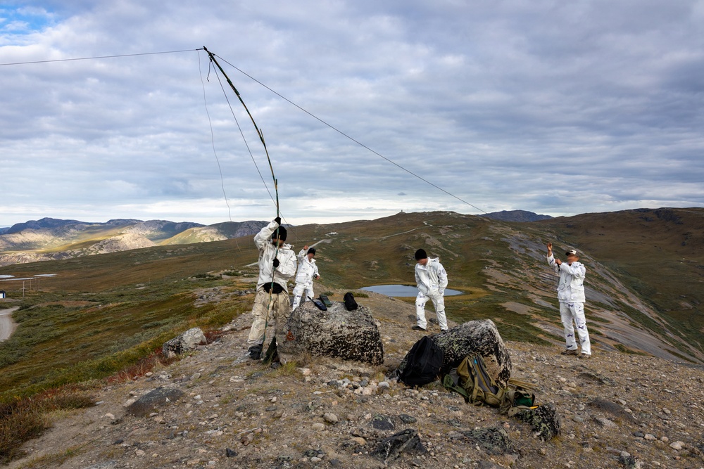 Four individuals wearing white camouflage overalls are setting up what appears to be a field radio antenna or wire antenna on a rocky hilltop in a tundra-like landscape. The terrain features rolling hills, sparse vegetation, and a small pond visible in the background. The activity suggests a military or paramilitary field communications exercise in a subarctic or arctic environment.