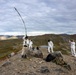 Four individuals wearing white camouflage overalls are setting up what appears to be a field radio antenna or wire antenna on a rocky hilltop in a tundra-like landscape. The terrain features rolling hills, sparse vegetation, and a small pond visible in the background. The activity suggests a military or paramilitary field communications exercise in a subarctic or arctic environment.