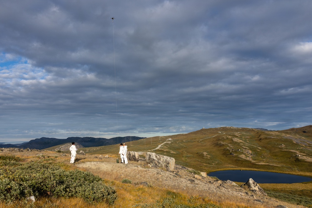 Two individuals dressed in white suits or coveralls stand on a rocky highland plateau, appearing to operate or observe a drone or kite flying high above them on a tether. The landscape features rolling alpine hills, sparse vegetation, a small dark lake, and distant structures on a ridgeline. The scene is set under a dramatic, partly cloudy sky with diffused sunlight illuminating the terrain.