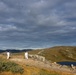 Two individuals dressed in white suits or coveralls stand on a rocky highland plateau, appearing to operate or observe a drone or kite flying high above them on a tether. The landscape features rolling alpine hills, sparse vegetation, a small dark lake, and distant structures on a ridgeline. The scene is set under a dramatic, partly cloudy sky with diffused sunlight illuminating the terrain.