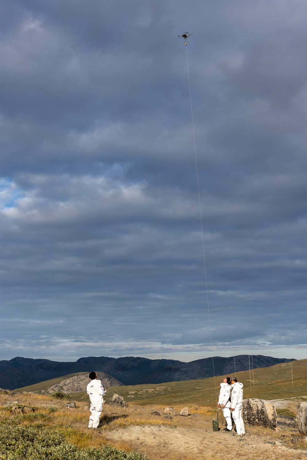 Three individuals wearing white full-body suits are operating a tethered drone in a remote, mountainous landscape under a dramatic cloudy sky. A visible tether line connects the drone high in the air to the ground team below. The setting appears to be an open highland or tundra environment with rocky terrain and sparse vegetation.