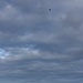 Three individuals wearing white full-body suits are operating a tethered drone in a remote, mountainous landscape under a dramatic cloudy sky. A visible tether line connects the drone high in the air to the ground team below. The setting appears to be an open highland or tundra environment with rocky terrain and sparse vegetation.