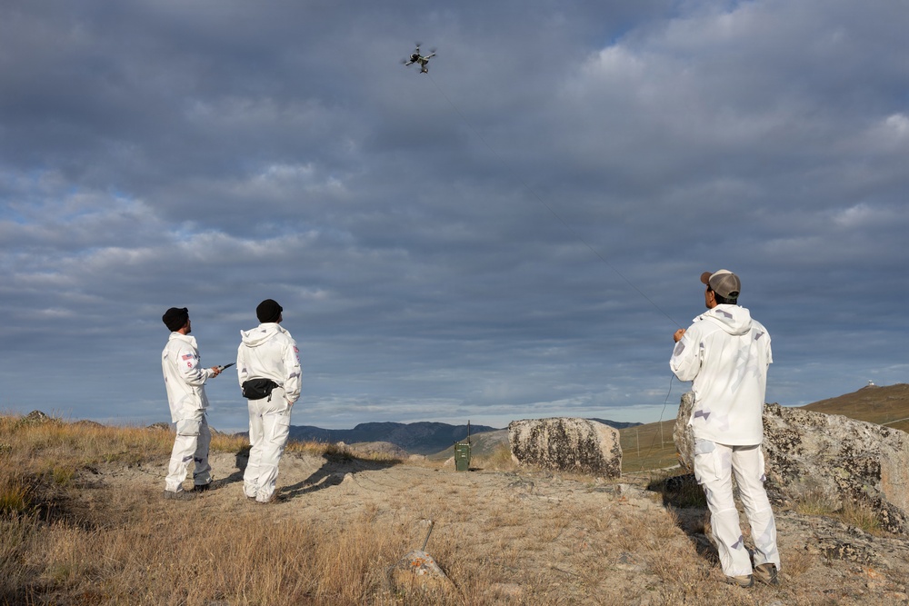 Three individuals dressed in white coveralls/jumpsuits are operating a quadcopter drone from an elevated rocky outcrop in a mountainous, arid landscape. One person on the left appears to be holding a remote controller while the others observe the drone in flight. The scene suggests a coordinated drone operation, possibly for surveying, research, or filming purposes.