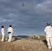 Three individuals dressed in white coveralls/jumpsuits are operating a quadcopter drone from an elevated rocky outcrop in a mountainous, arid landscape. One person on the left appears to be holding a remote controller while the others observe the drone in flight. The scene suggests a coordinated drone operation, possibly for surveying, research, or filming purposes.