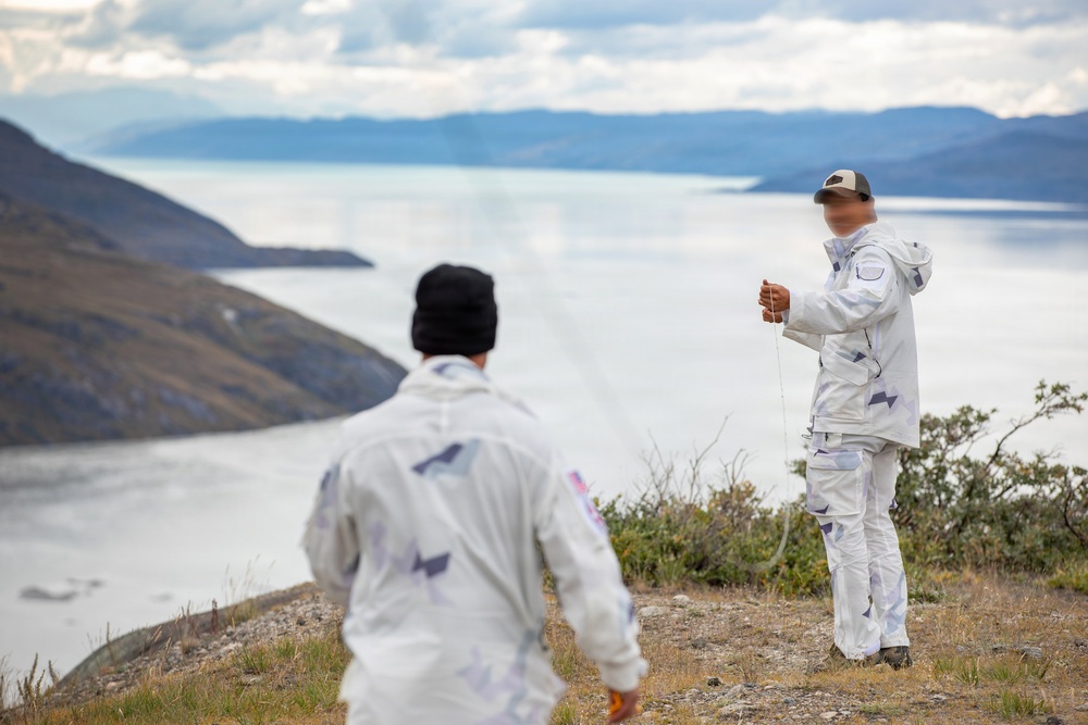 Two individuals wearing matching white camouflage-patterned suits stand on an elevated rocky terrain overlooking a large body of water with mountains in the background. The person in the foreground faces away from the camera wearing a black beanie, while the person in the background faces forward wearing a cap and appears to be holding a thin cord or line. The landscape suggests a remote, cold-weather environment, possibly Patagonia or a similar sub-arctic region.