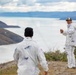 Two individuals wearing matching white camouflage-patterned suits stand on an elevated rocky terrain overlooking a large body of water with mountains in the background. The person in the foreground faces away from the camera wearing a black beanie, while the person in the background faces forward wearing a cap and appears to be holding a thin cord or line. The landscape suggests a remote, cold-weather environment, possibly Patagonia or a similar sub-arctic region.