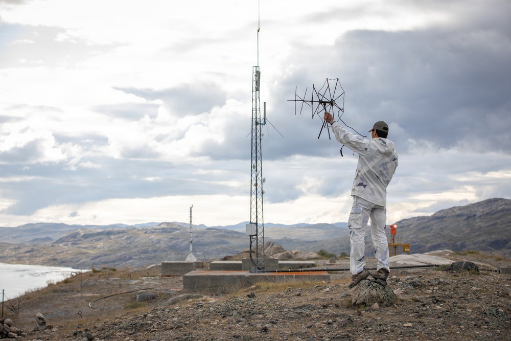 A person dressed in white work clothing and a cap stands on a rocky hilltop, holding up what appears to be a directional antenna or signal-receiving device. In the background, a telecommunications tower and associated ground equipment are visible, set against a mountainous landscape with overcast skies. The setting appears to be a remote, arctic or sub-arctic region based on the terrain and vegetation.