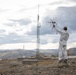 A person dressed in white work clothing and a cap stands on a rocky hilltop, holding up what appears to be a directional antenna or signal-receiving device. In the background, a telecommunications tower and associated ground equipment are visible, set against a mountainous landscape with overcast skies. The setting appears to be a remote, arctic or sub-arctic region based on the terrain and vegetation.