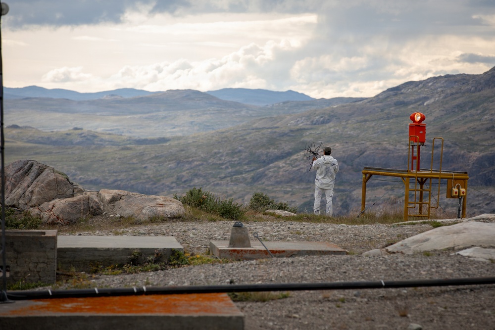 A person dressed in white work clothing stands on a rocky elevated plateau holding what appears to be a small antenna or directional equipment, facing a vast mountainous landscape. To their right is a yellow metal safety railing structure with a red beacon or warning light mounted on top. The setting appears to be a remote industrial or monitoring installation situated in rugged, treeless highland terrain.
