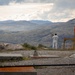 A person dressed in white work clothing stands on a rocky elevated plateau holding what appears to be a small antenna or directional equipment, facing a vast mountainous landscape. To their right is a yellow metal safety railing structure with a red beacon or warning light mounted on top. The setting appears to be a remote industrial or monitoring installation situated in rugged, treeless highland terrain.