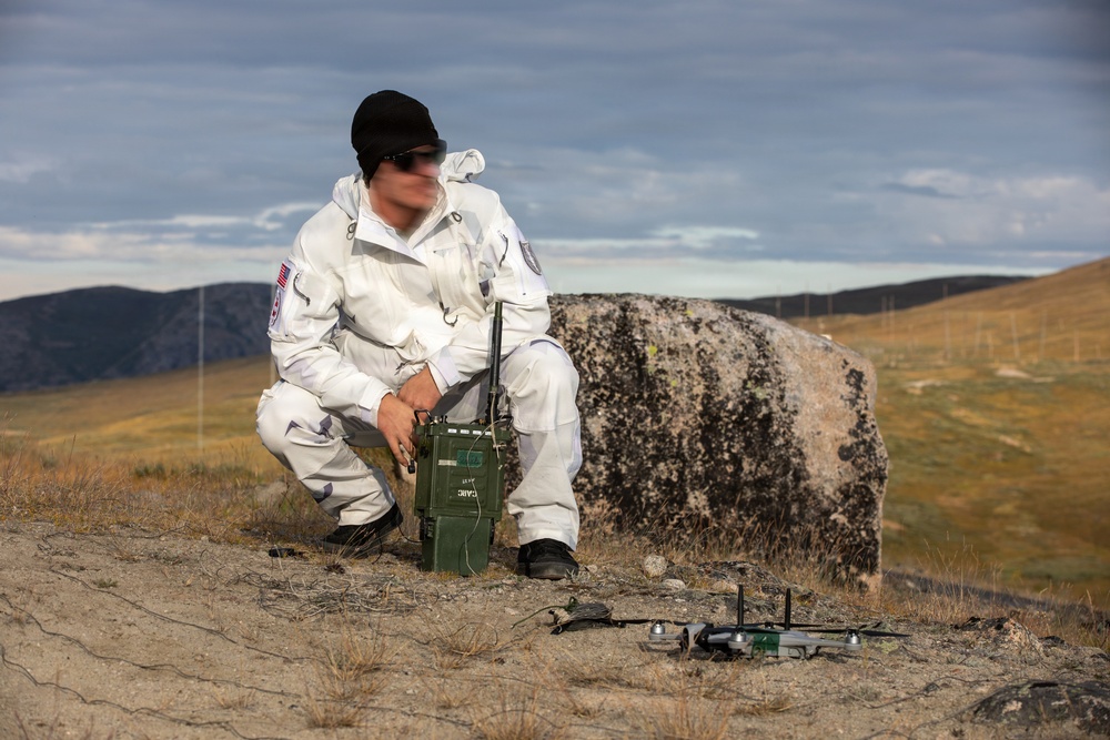 A person wearing a white uniform with a US flag patch and black beanie is crouching outdoors in a rugged, open landscape. They are operating what appears to be a military-style green radio or communications device, with a small quadcopter drone placed on the ground nearby. The setting appears to be a remote, arid terrain with rocky outcroppings and hills in the background.