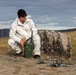 A person wearing a white uniform with a US flag patch and black beanie is crouching outdoors in a rugged, open landscape. They are operating what appears to be a military-style green radio or communications device, with a small quadcopter drone placed on the ground nearby. The setting appears to be a remote, arid terrain with rocky outcroppings and hills in the background.